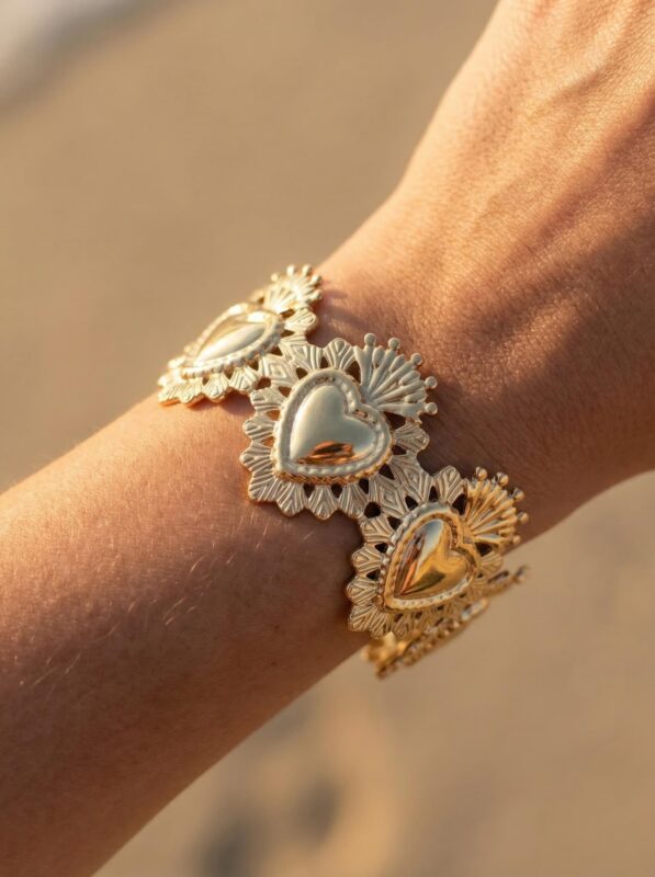 Bracelet manchette cœur sacré doré, jonc manchette sacré coeur en acier inoxydable doré porté au poignet, motifs baroques ciselés en relief, photographié en lumière naturelle dorée sur fond de sable